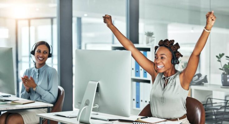 A woman sitting at her desk, raising her arms in the air and smiling. There's a woman at another desk smiling and clapping.