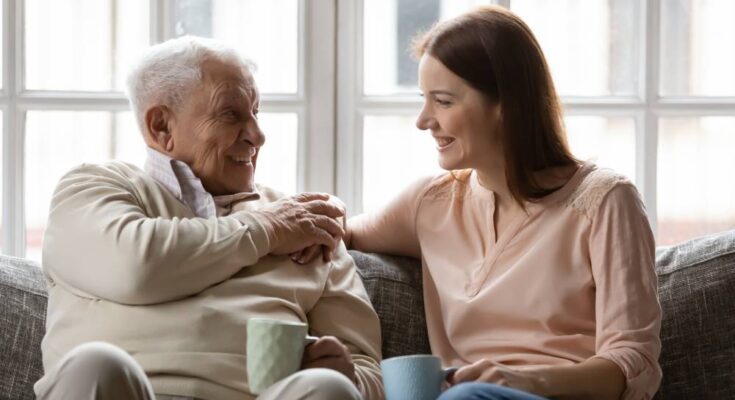 An old father and his young-adult daughter sit close together on a couch, holding mugs and smiling at each other.