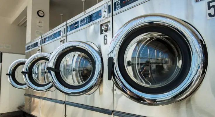 A row of numbered stainless steel commercial washing machines sit with their doors open in a laundromat.