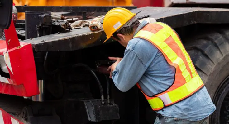 A heavy equipment engineer wearing a hard hat and safety vest uses grease to lubricate the axle of a crane.