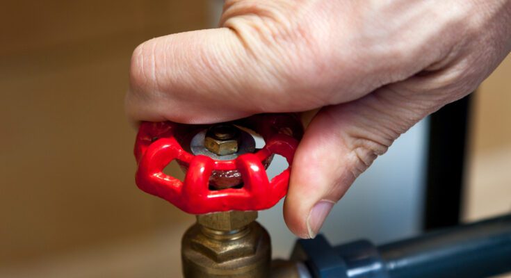 A close-up view shows a hand on the red knob of a brass water valve that links two blue water pipes.