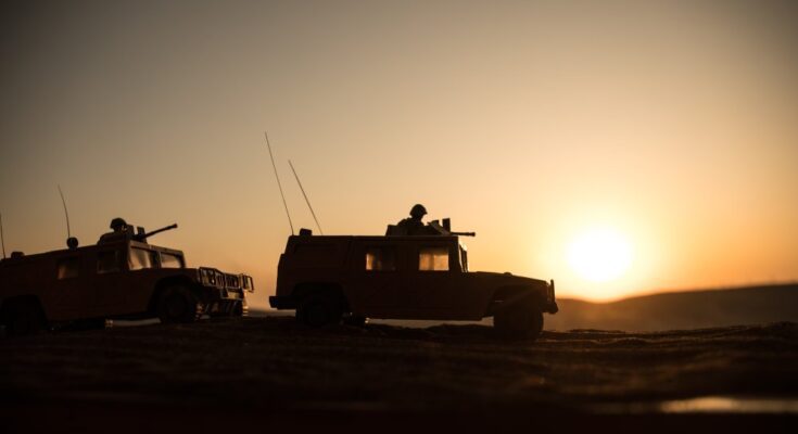 At sunset, two military vehicles, each manned by a soldier, navigate rough, muddy terrain under a golden sky.