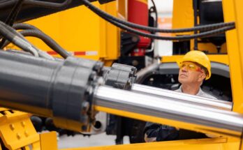 A construction worker in a yellow hard hat examines the clean hydraulic cylinder on a yellow excavator.