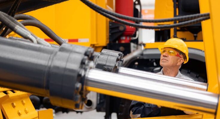 A construction worker in a yellow hard hat examines the clean hydraulic cylinder on a yellow excavator.