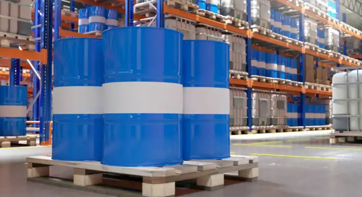 A warehouse with dozens of blue and white barrels and plastic containers placed on pallets and high metal shelves.