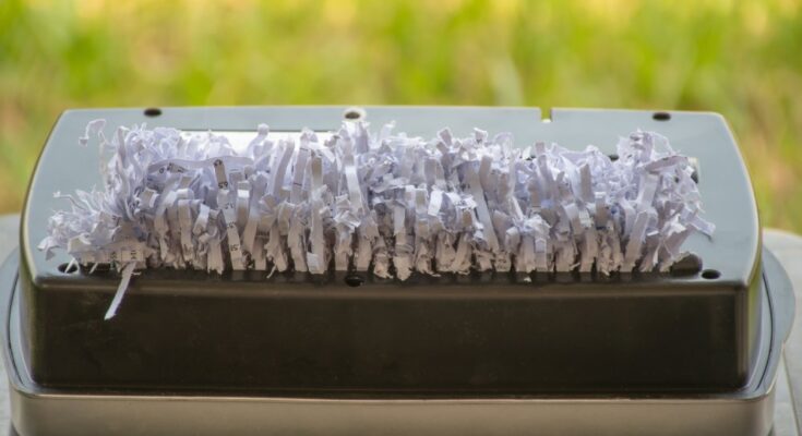 A paper shredder with a jam full of white paper shreds. The paper shredder is near a window that shows grass.