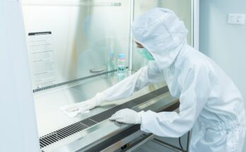 A scientist wears a sterile coverall gown and wipes down a metal surface in a cleanroom.