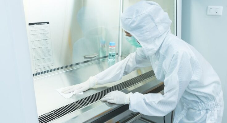 A scientist wears a sterile coverall gown and wipes down a metal surface in a cleanroom.