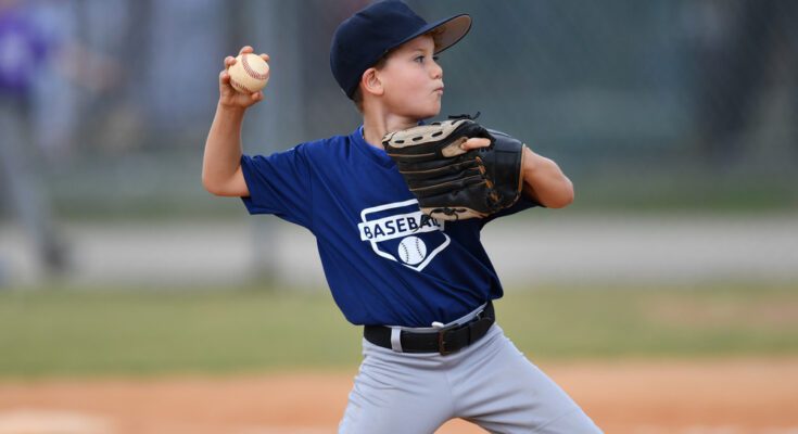 A young baseball athlete pitching in a game. He's mid-throw, wearing gray pants and a navy blue uniform top.