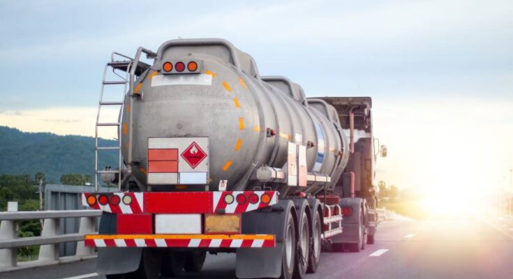 A large industrial truck driving on a road transporting flammable chemicals in a large cylindrical metal tank.