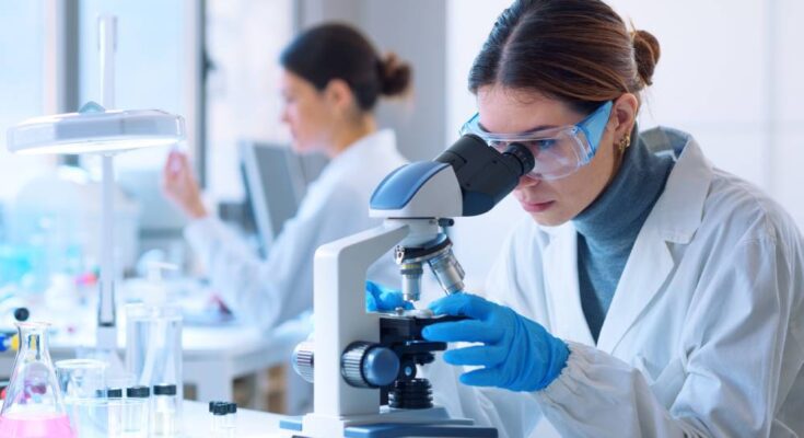 A woman in a lab coat, gloves, and goggles looks at a microscope. Another woman works behind her.