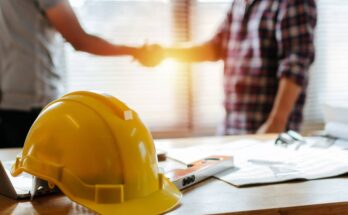 The sun shines through a window onto two people shaking hands next to a table with a yellow hard hat on it.