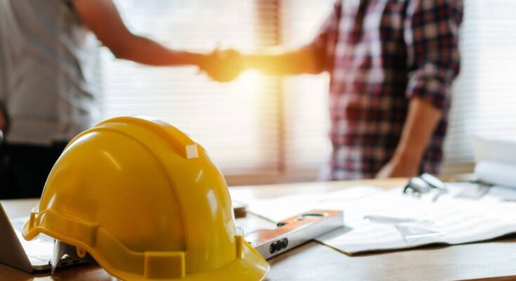 The sun shines through a window onto two people shaking hands next to a table with a yellow hard hat on it.