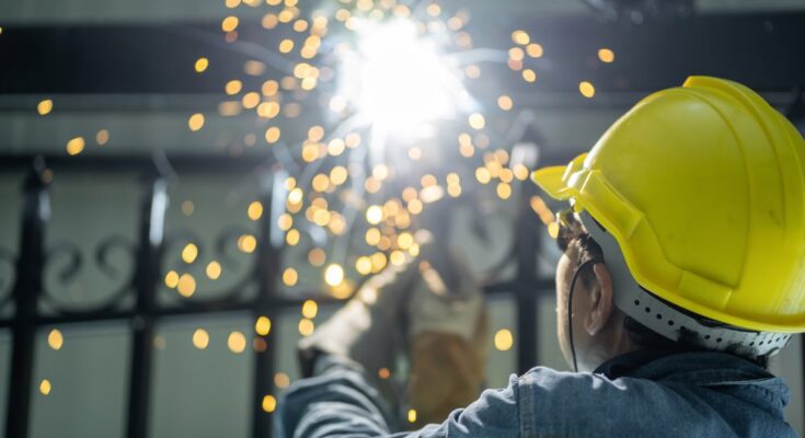 A man wearing a yellow hard hat and denim jacket while grinding steel. Sparks are flying around in front of his face.