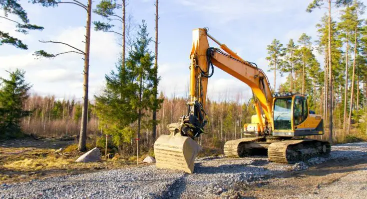 An excavator on a property near the woods. There's gravel on the ground and a tree line behind the excavator.