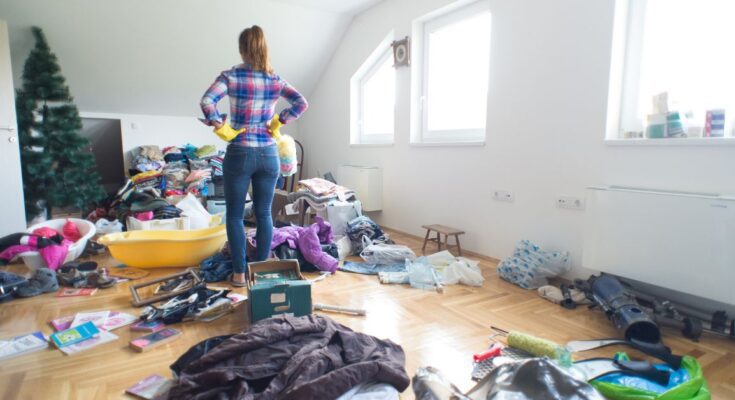 A woman stands ready to clean a messy room. She's wearing yellow gloves and has her hands on her hips.