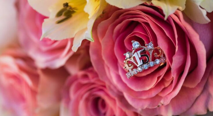 A silver ring shaped like a tiara with the number "15" at its center displayed in a bouquet of roses and lilies.