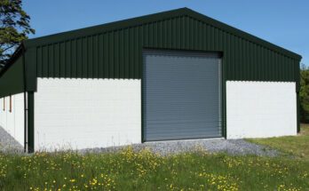 The exterior of a green and white metal barn with a peaked roof and a large gray door. The barn is surrounded by trees.