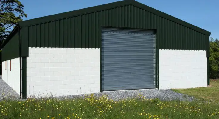 The exterior of a green and white metal barn with a peaked roof and a large gray door. The barn is surrounded by trees.