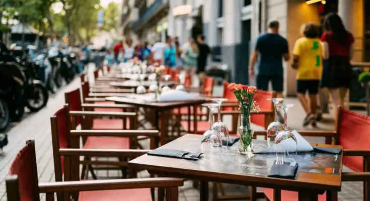 Wooden tables and chairs with red fabric line the sidewalk outside a cafe. There are wine glasses and flowers on the tables.