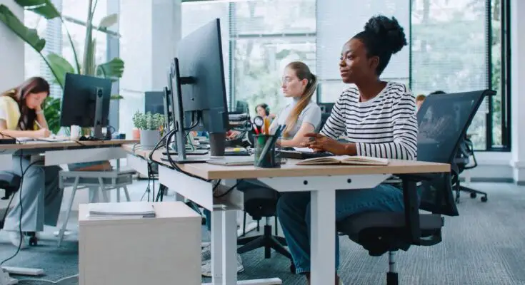 Three women sitting at desks with large monitors and keyboards. The office has tall windows with open blinds.