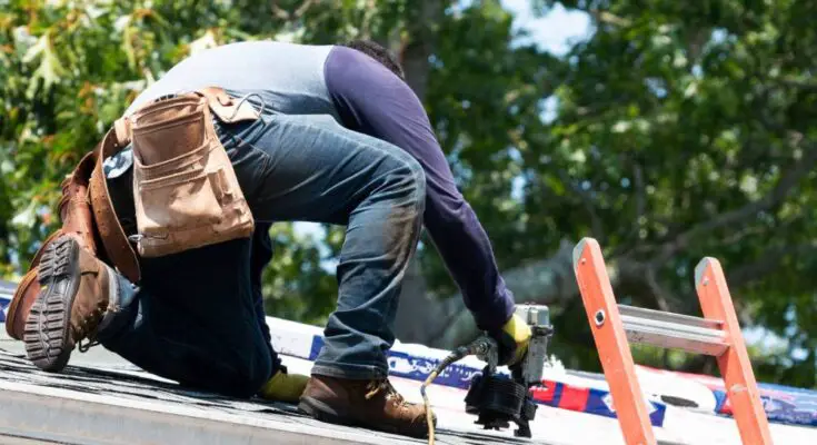 Worker wearing a tool belt using a nail gun on the roof of a home next to an orange ladder with trees in background.