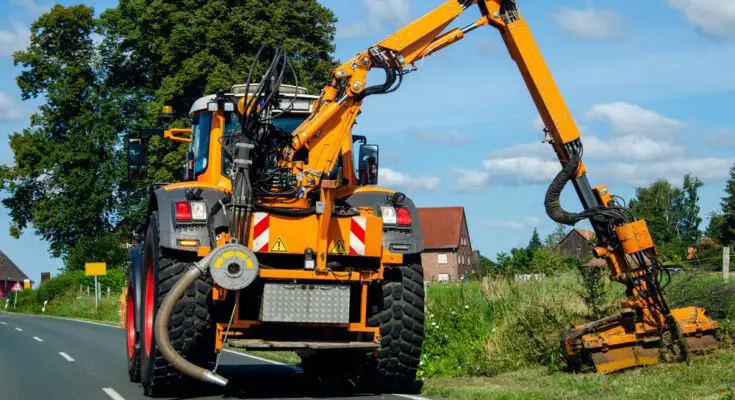 A large, yellow tractor drives down a road while using a boom mower to mow the overgrown vegetation.