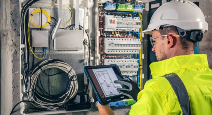 An electrical worker wearing a reflective uniform and a white hard hat is holding a tablet in front of a switchboard.