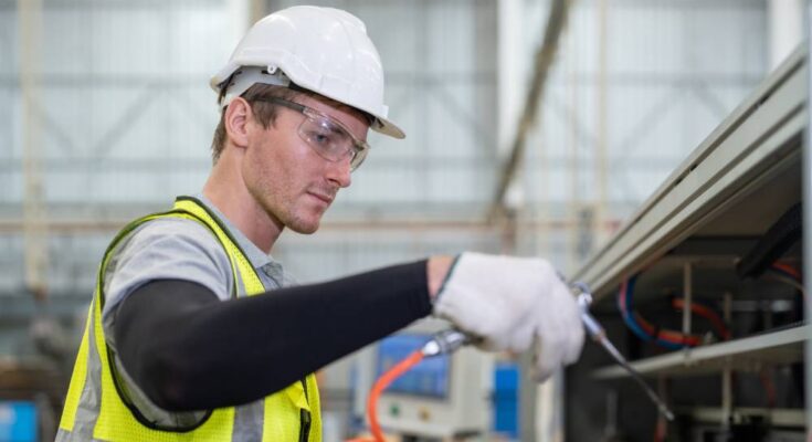 A male engineer with safety goggles cleans a packaging machine using an air duster in a factory warehouse.