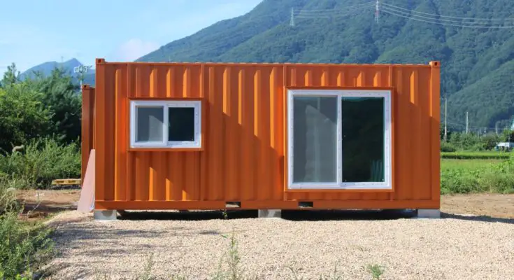 An orange shipping container building with a siding glass door and a window sits in a gravel lot near mountains.