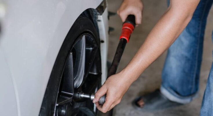 A close-up of a person in jeans and outdoor sandals carefully tightening the lug nuts on a car's wheel.