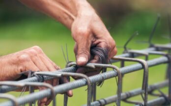 A close-up of a man's hands tying wire around the bent joints of metal bars to attach them to rebar.