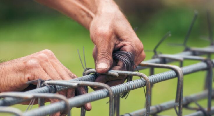 A close-up of a man's hands tying wire around the bent joints of metal bars to attach them to rebar.