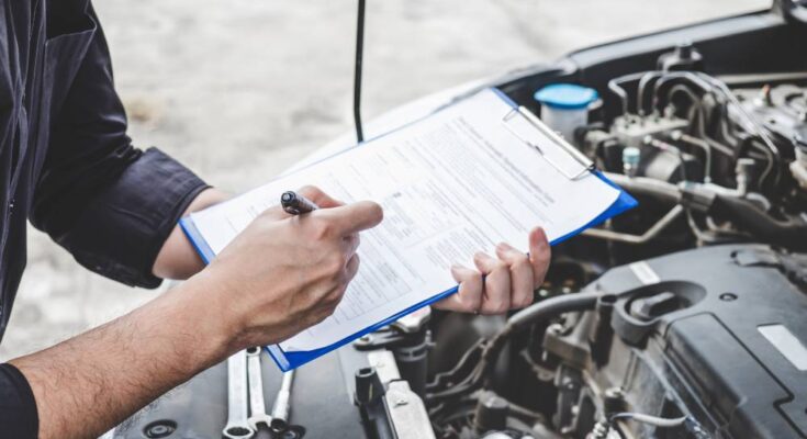 A person looking underneath the hood of a car. They hold a clipboard and a pen to make notes on a document.