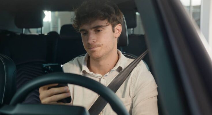 A frustrated looking young man sits in the driver's seat of a vehicle with one hand holding his phone.