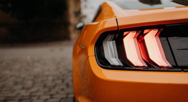 The left rear brake light of an orange Ford Mustang is visible from behind. The light is illuminated and red.