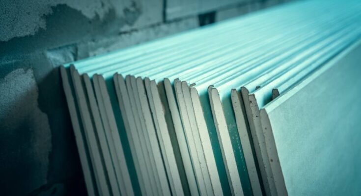 A close-up of a stack of green moisture-resistant drywall sheets leaning up against a brick wall.