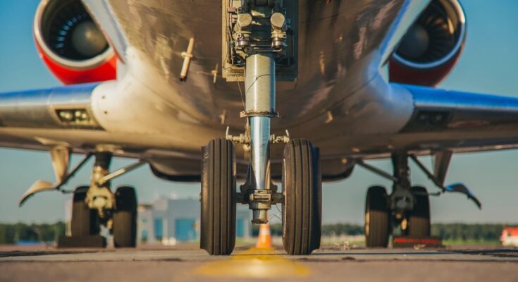 Large chucks hold a plane by its landing gear in place at an airport runway. Buildings are visible in the distance.