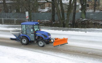 A compact blue tractor using a front-mounted snow plow attachment and rear-mounted salt spreader to clear a road.