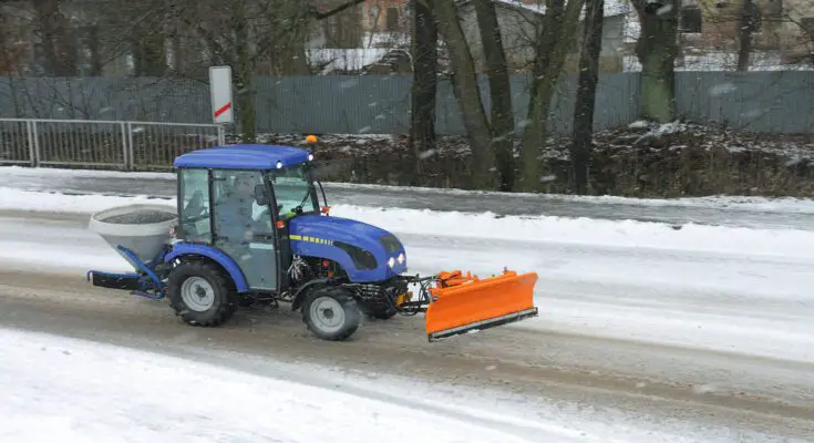 A compact blue tractor using a front-mounted snow plow attachment and rear-mounted salt spreader to clear a road.