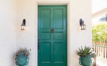 A white home with a shaded front entryway. The front door is teal and there's two matching teal planters on either side.