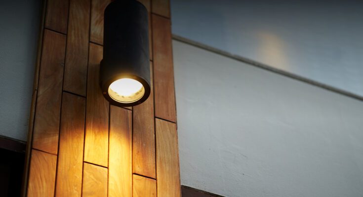 A decorative black wall sconce attached to a section of wood paneling on a wall. The fixture is producing soft accent lighting.