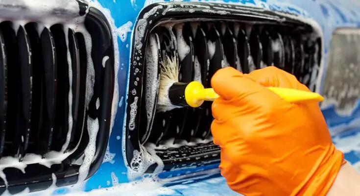 A person with an orange glove using a boar's hair brush to clean and detail the sudsy grill of a blue vehicle.