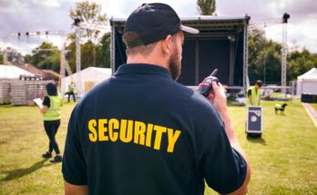 A man wearing a blue shirt that says "Security" standing in front of an outdoor stage while holding a two-way radio.