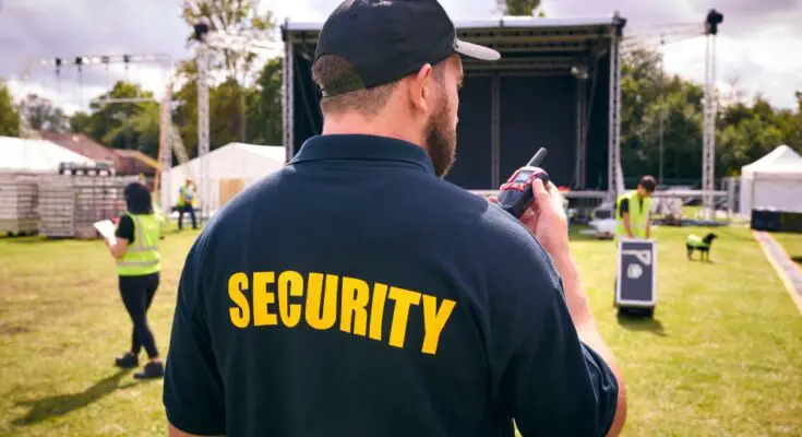 A man wearing a blue shirt that says "Security" standing in front of an outdoor stage while holding a two-way radio.