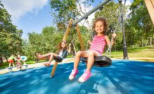 Two little girls are swinging on a swing with a black seat and chain links. The flooring is a blue rubber.