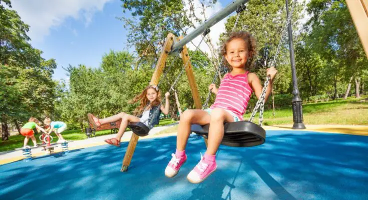 Two little girls are swinging on a swing with a black seat and chain links. The flooring is a blue rubber.