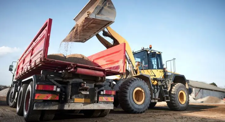 A yellow excavator lifts up a pile of dirt to dump in the back of an orange-colored dump truck at a construction site.