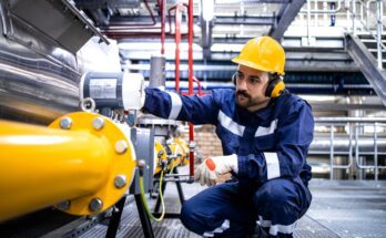 A man in a jumpsuit and yellow hardhat kneels down to examine a yellow pipe in an industrial plant.