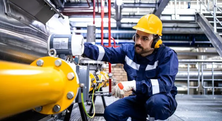 A man in a jumpsuit and yellow hardhat kneels down to examine a yellow pipe in an industrial plant.